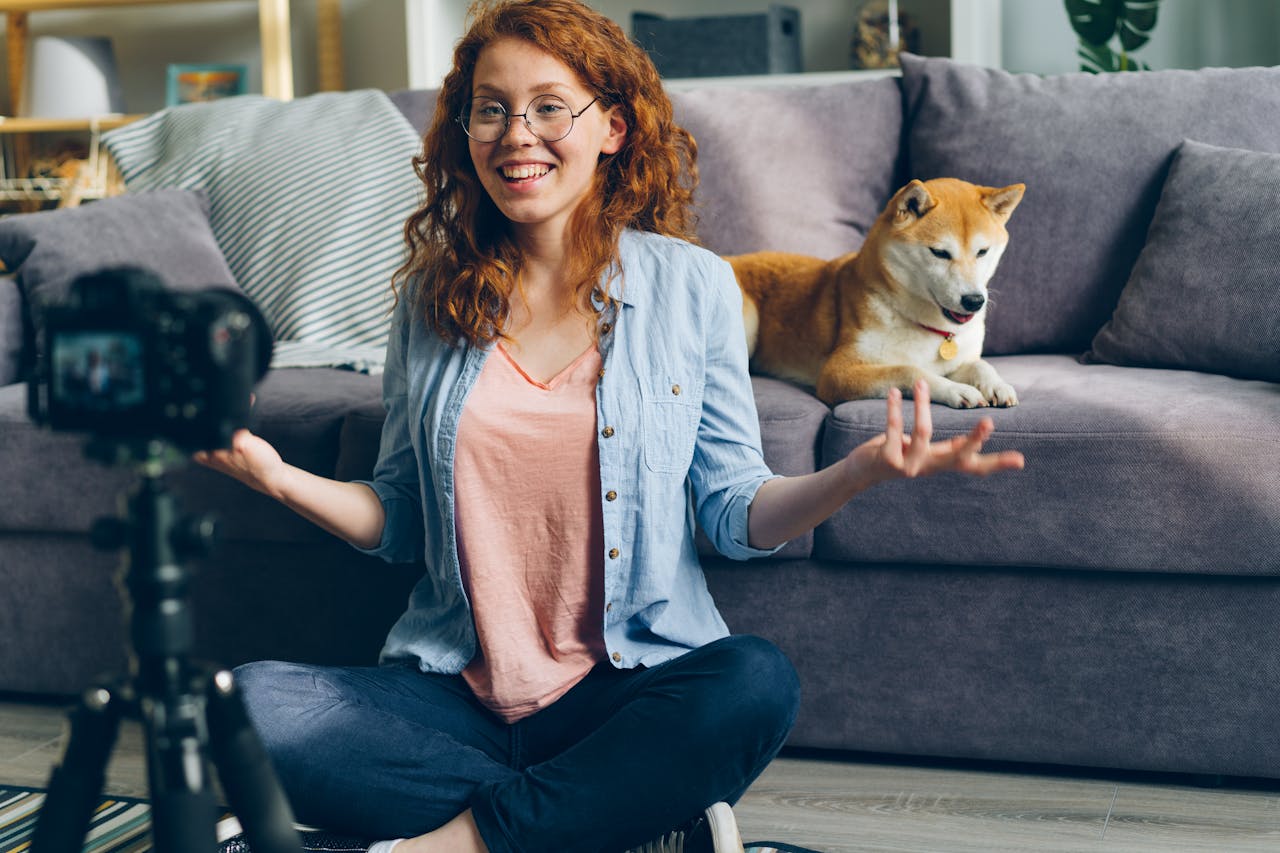 A smiling woman records a vlog with her Shiba Inu dog on a couch in a cozy living room setting.