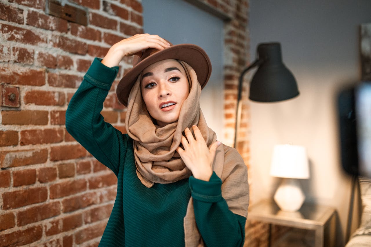 Portrait of a Muslim woman wearing a hijab and hat against a chic indoor brick backdrop.